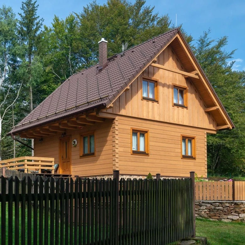 Dřevěný roubenkový dům s tmavou střechou před lesním pozadím.