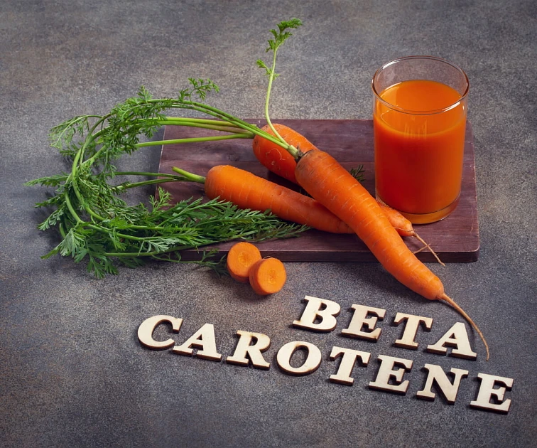 Čerstvé mrkve s listy, šťáva a dřevěné písmena 'BETA CAROTENE' na tmavém povrchu.