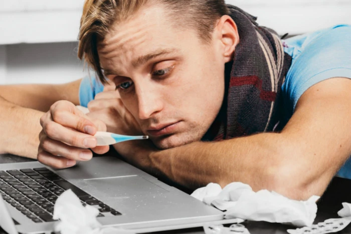 Muž s teploměrem u úst leží na laptopu při angíně.