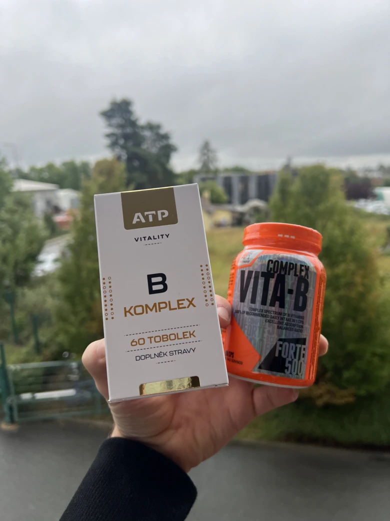Ruka drží balíček vitamínu B komplexu vedle oranžového flakonu.