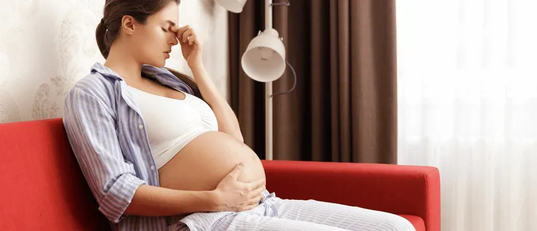 Těhotná žena sedí na gauči a drží si hlavu kvůli bolesti hlavy.