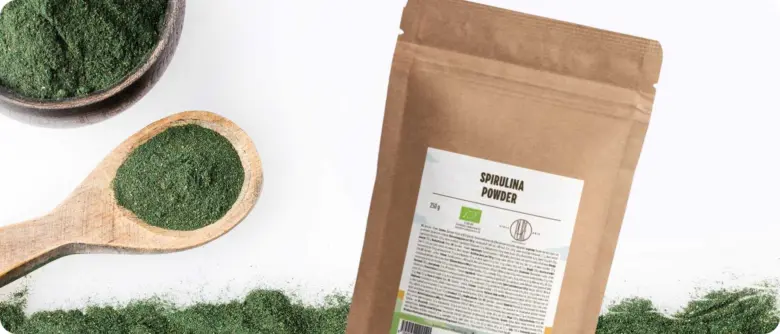 Balíček s práškem z řasy Spirulina vedle lžičky plné zeleného prášku.