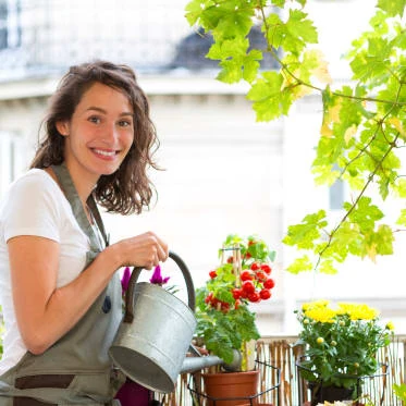 Žena zalévá kvetoucí rostliny na balkóně v městském prostředí.