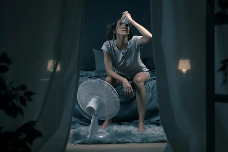 Žena sedí na posteli v noci s ventilátorem kvůli nočnímu pocení.