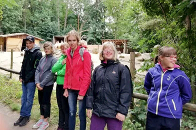 Skupina lidí venku v přírodě u dřevěných staveb.