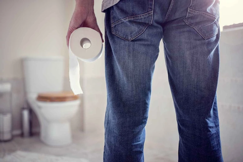 Muž drží toaletní papír v koupelně s WC na pozadí.