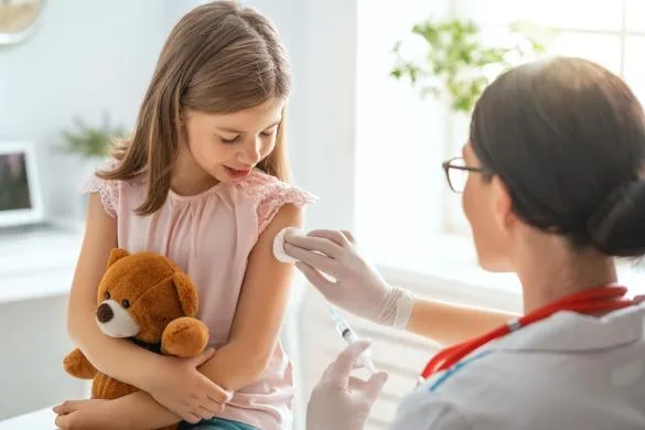 Dívka dostává očkování od sestry s medvídkem v náručí.