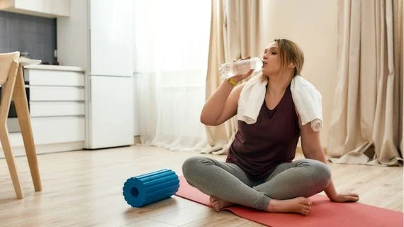 Žena pije vodu po cvičení s válečkem na podlaze.