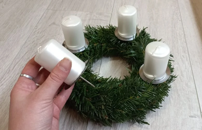 Ruka zasouvá bílou svíčku do zeleného adventního věnce.